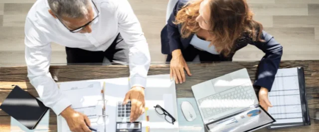 Zwei Personen am Schreibtisch mit Laptop. Dokumenten und Taschenrechner