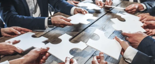 Mehrere Personen an einem Tisch mit jeweils einem großen Puzzleteil in der Hand