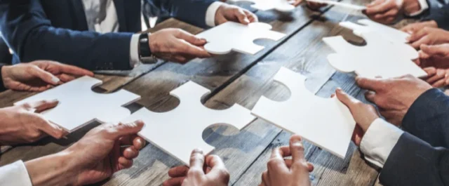 mehrere Personen in Businesskleidung halten große weiße Puzzlestücke zusammen